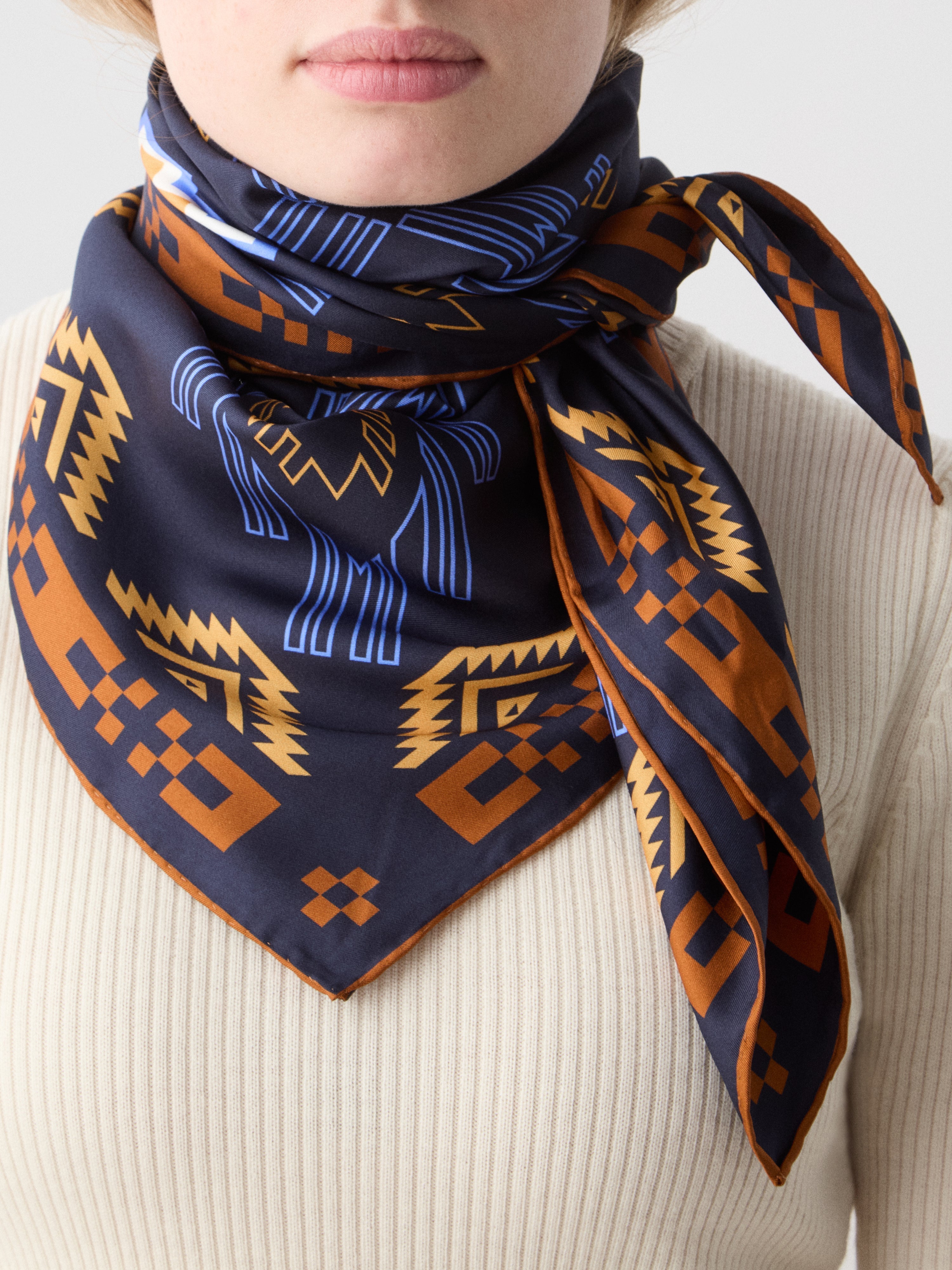 We Norwegians | FW25 | Pioneer Silk Scarf_Navy blue | Female model wearing the scarf with printed motifs over a light top | Shot against an off-white background