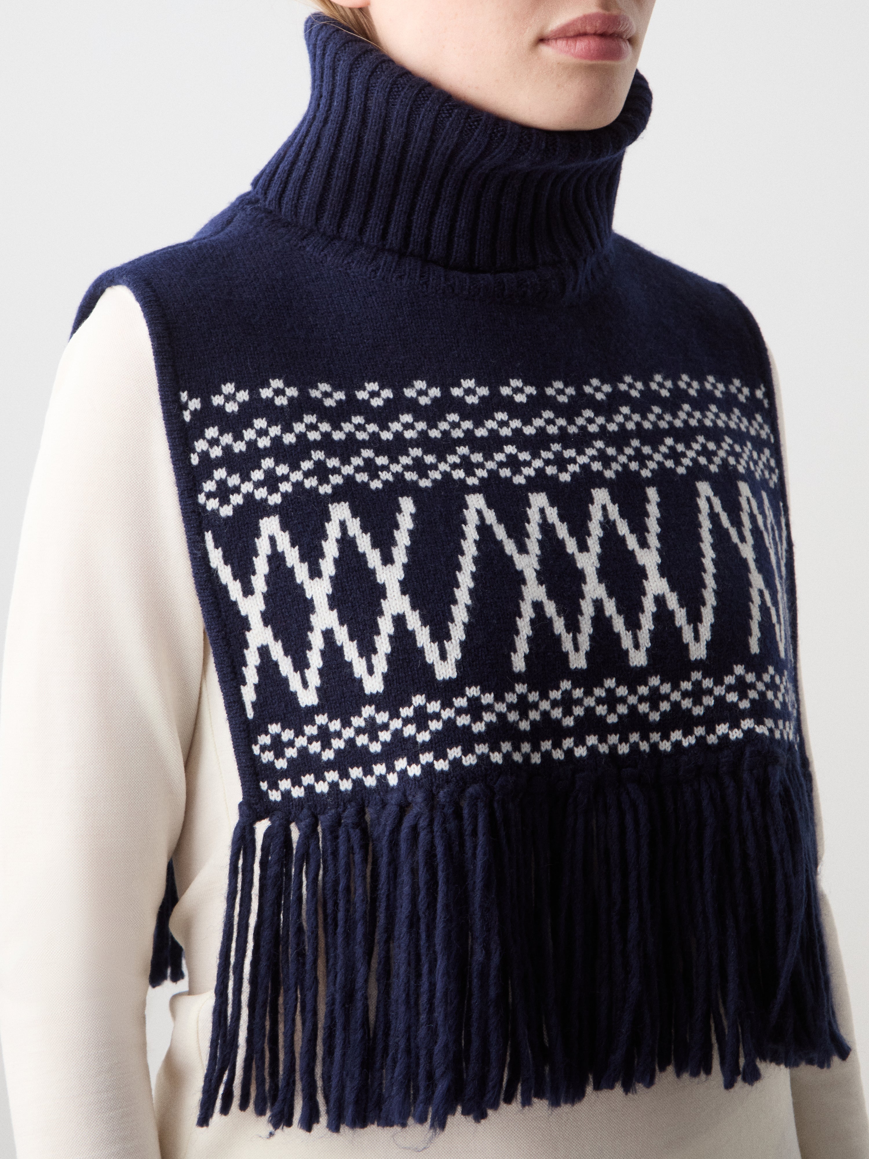 We Norwegians | FW25 | Setesdal Bib_Navy blue | Model is wearing the knitted bib with a turtle neck and fine fringes at the hem over a white sweater, against an off-white background